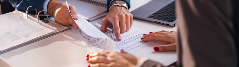 Three people review and discuss documents at a desk. One person points at a paper, another holds it, and the third has hands resting on the table. An open binder and a laptop are also visible.