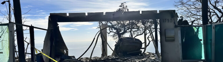 A burned, roofless building with broken glass and charred debris stands overlooking the ocean under a bright sky. Yellow caution tape is visible on the left; leafless trees surround the structure.