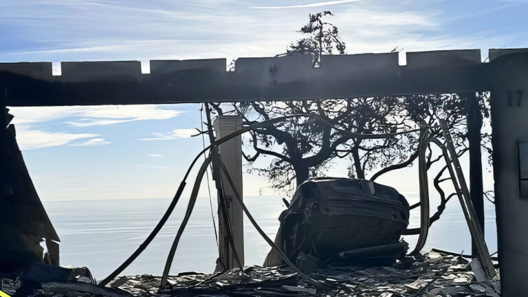 A burned, roofless building with broken glass and charred debris stands overlooking the ocean under a bright sky. Yellow caution tape is visible on the left; leafless trees surround the structure.