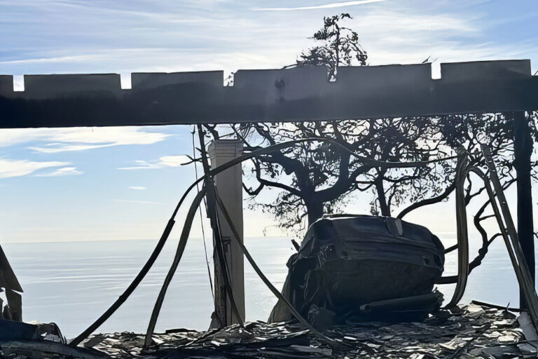 A burned, roofless building with broken glass and charred debris stands overlooking the ocean under a bright sky. Yellow caution tape is visible on the left; leafless trees surround the structure.