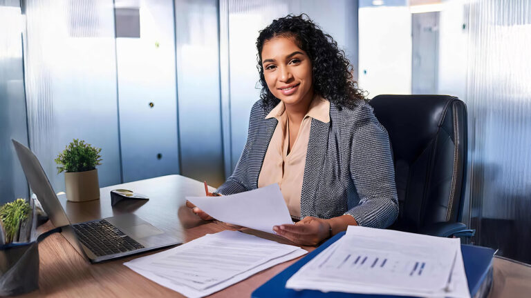 A woman in business attire sits at a desk in a modern office, smiling while holding papers. A laptop, documents, and potted plants are on the desk. Glass walls and office decor are visible in the background.