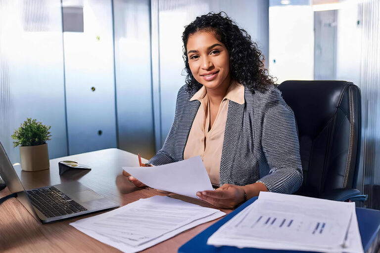A woman in business attire sits at a desk in a modern office, smiling while holding papers. A laptop, documents, and potted plants are on the desk. Glass walls and office decor are visible in the background.