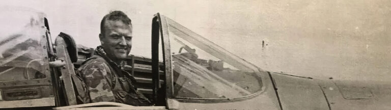 A black-and-white photo of a smiling pilot in military gear sitting in the cockpit of a fighter jet, with the plane canopy open.