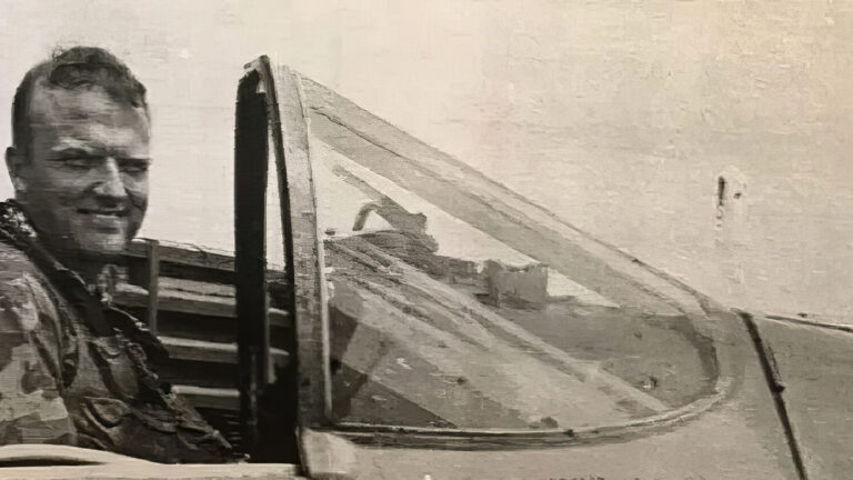 A black-and-white photo of a smiling pilot in military gear sitting in the cockpit of a fighter jet, with the plane canopy open.