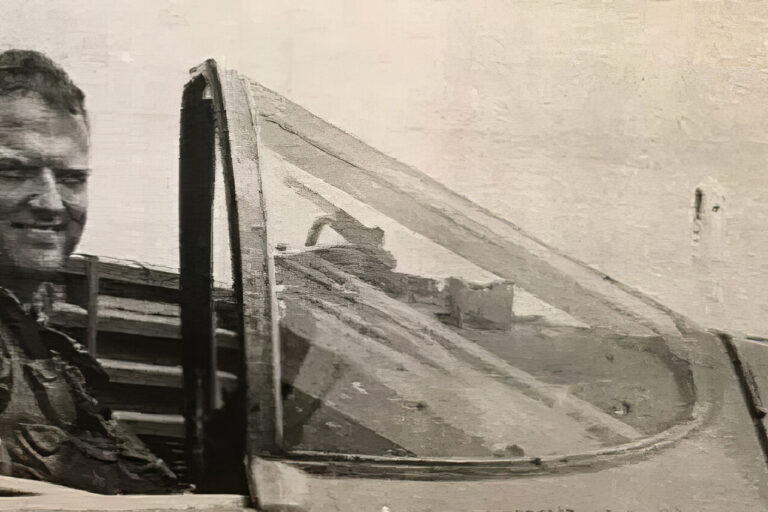 A black-and-white photo of a smiling pilot in military gear sitting in the cockpit of a fighter jet, with the plane canopy open.