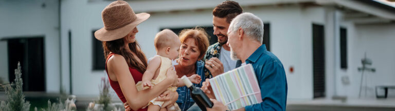 A group of adults smiling and gathered around a baby outdoors. One person is holding a wrapped gift, and another is holding the baby, who is wearing a sun hat. A white house is visible in the background.