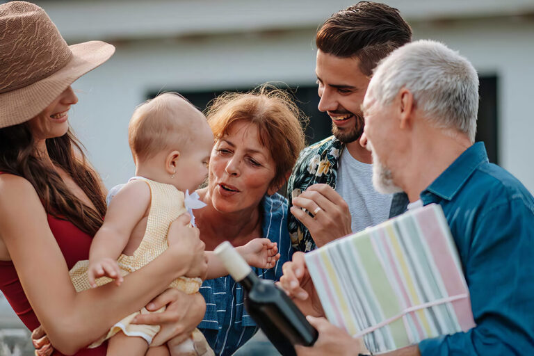 A group of adults smiling and gathered around a baby outdoors. One person is holding a wrapped gift, and another is holding the baby, who is wearing a sun hat. A white house is visible in the background.