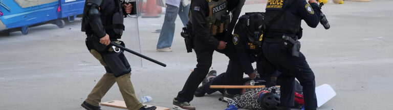 Police officers in riot gear detain and restrain a person on the ground during a tense situation outdoors, with batons and equipment scattered nearby. Other people are visible in the background.