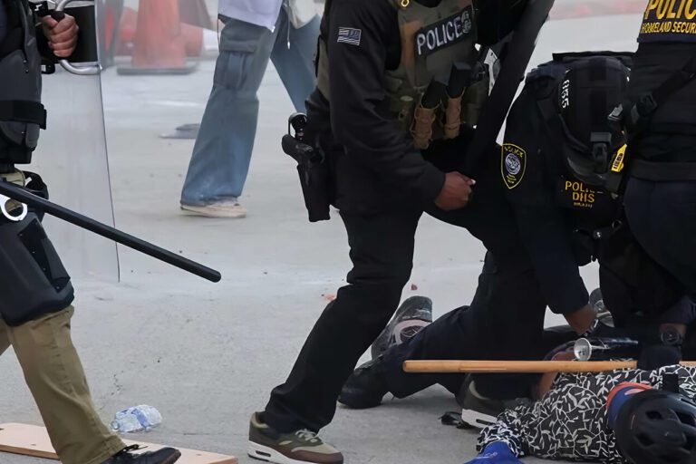 Police officers in riot gear detain and restrain a person on the ground during a tense situation outdoors, with batons and equipment scattered nearby. Other people are visible in the background.
