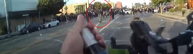 A person holds a weapon aimed at a large crowd in an urban street; many people are running or gathered in the distance. The scene appears tense and chaotic. A red circle highlights a figure in the crowd.