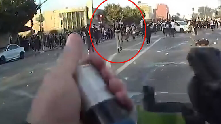 A person holds a weapon aimed at a large crowd in an urban street; many people are running or gathered in the distance. The scene appears tense and chaotic. A red circle highlights a figure in the crowd.
