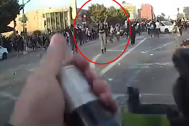 A person holds a weapon aimed at a large crowd in an urban street; many people are running or gathered in the distance. The scene appears tense and chaotic. A red circle highlights a figure in the crowd.