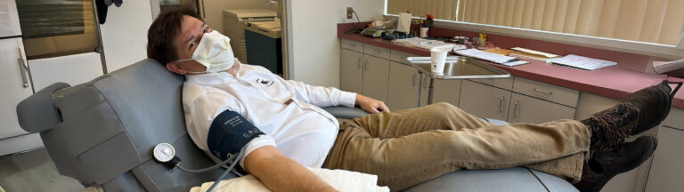 A man wearing a mask reclines on a medical chair while donating blood. He has a blood pressure cuff on his arm and is in a clinic room with cabinets, papers, and medical equipment visible in the background.