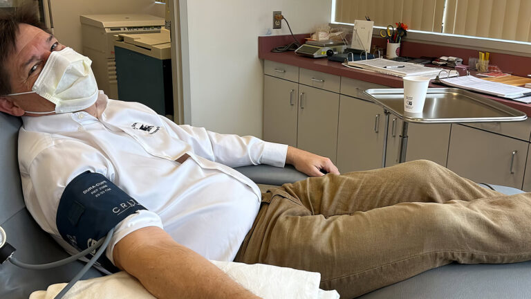 A man wearing a mask reclines on a medical chair while donating blood. He has a blood pressure cuff on his arm and is in a clinic room with cabinets, papers, and medical equipment visible in the background.