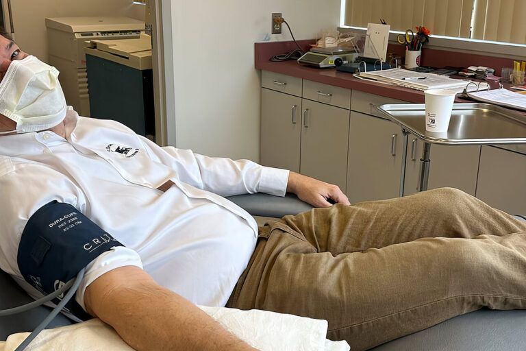 A man wearing a mask reclines on a medical chair while donating blood. He has a blood pressure cuff on his arm and is in a clinic room with cabinets, papers, and medical equipment visible in the background.