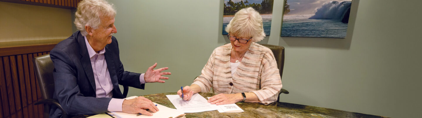 Two older adults in business attire sit at a desk; the woman signs a document with a pen while the man gestures and smiles. Framed ocean photos hang on the wall behind them.