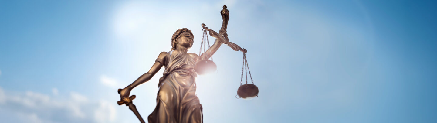 A bronze statue of Lady Justice holding balanced scales and a sword, blindfolded, stands against a bright blue sky with sunlight shining beside her.
