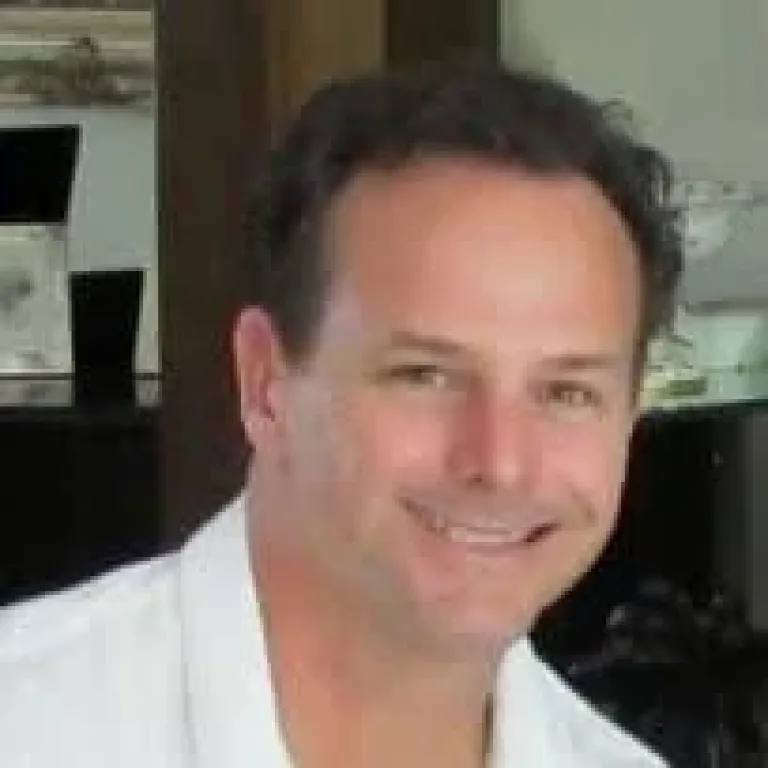 A man with short dark hair smiles at the camera, wearing a white shirt. The background shows a blurred indoor setting with framed pictures and decor.