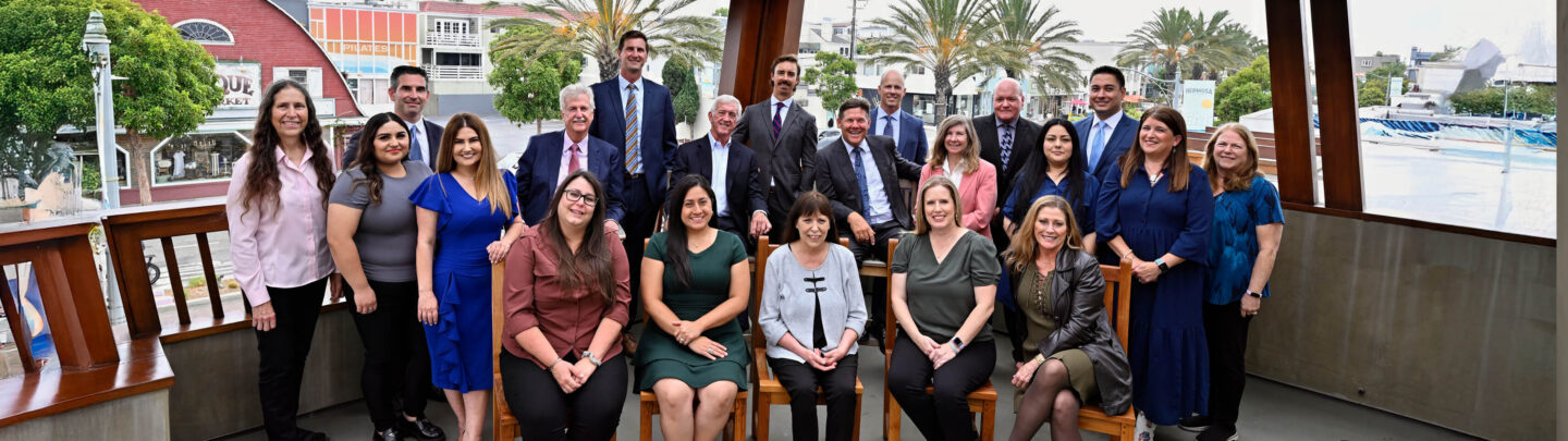 A group of 21 professionally dressed people pose and smile together on a covered outdoor deck, with trees, buildings, and palm trees visible in the background.