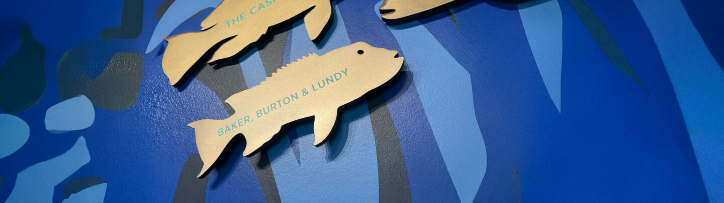Wooden fish-shaped plaques with names, including BAKER, BURTON & LUNDY, are mounted on a blue wall featuring abstract aquatic patterns.