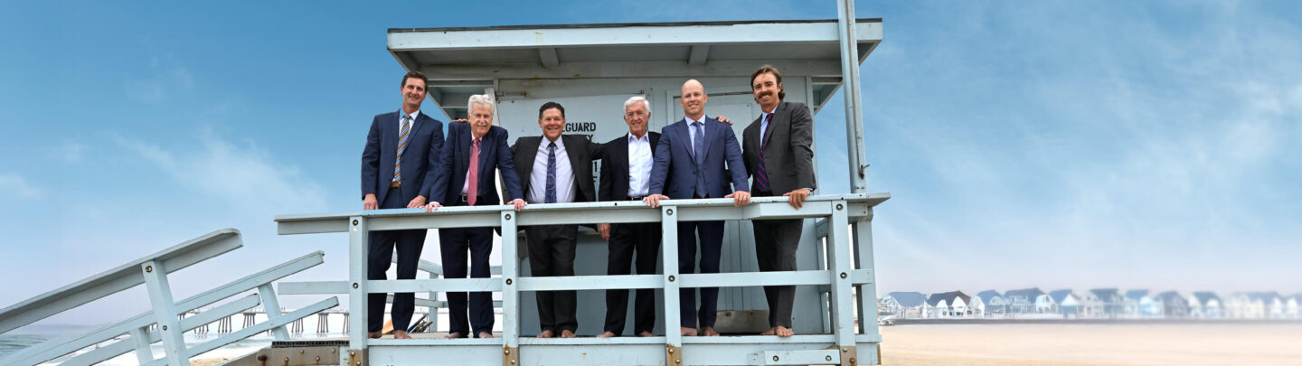 Six men in suits stand barefoot on a lifeguard tower at the beach, smiling at the camera with houses and a clear blue sky in the background.
