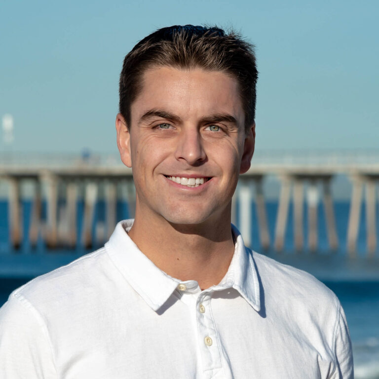 A young man with short brown hair, wearing a white collared shirt, smiles at the camera. In the background, there is a blurred pier and blue water under a clear sky.