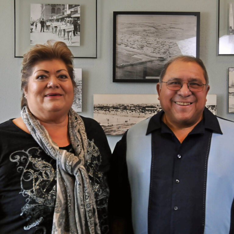 Two adults stand side by side smiling, in front of a wall decorated with black and white photos. The woman wears a patterned scarf and the man wears glasses and a collared shirt.