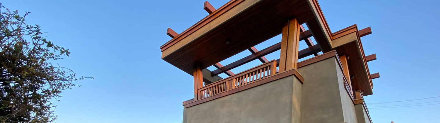 A modern two-story building with stone and concrete exterior, wooden accents, large windows, and balcony, set on a paved sidewalk with benches, plants, and clear blue sky in the background.
