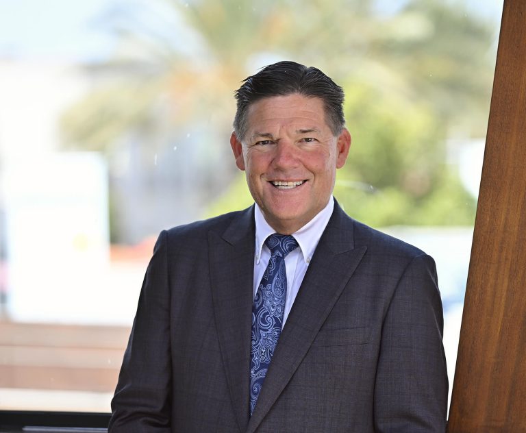 A man in a dark suit and blue patterned tie smiles while standing indoors by a large window, with blurred greenery and a palm tree visible outside in the background.