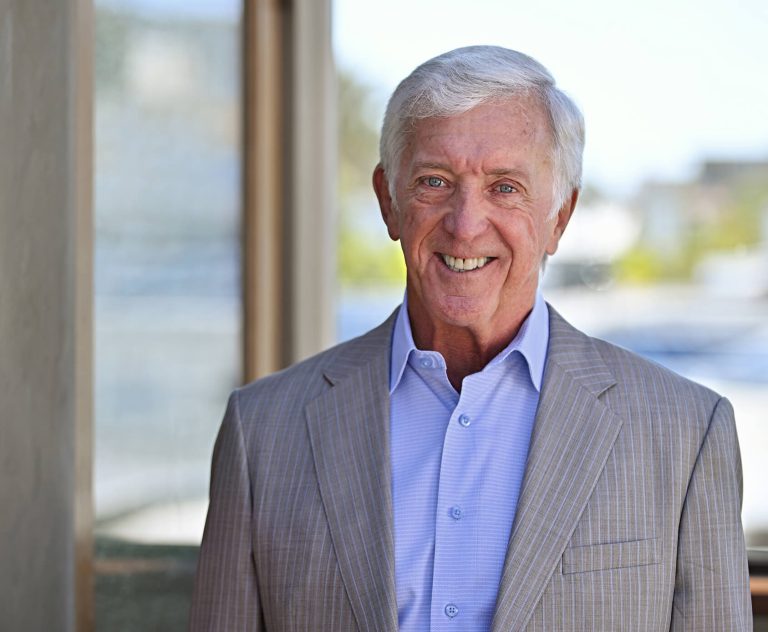An older man with white hair smiles warmly, wearing a light gray pinstripe suit jacket over a light blue collared shirt, standing in front of a softly blurred outdoor background.