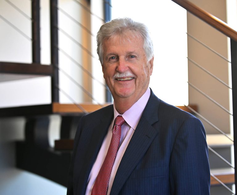 An older man in a dark suit, light pink shirt, and red patterned tie stands smiling indoors in front of a modern staircase with metal railings.