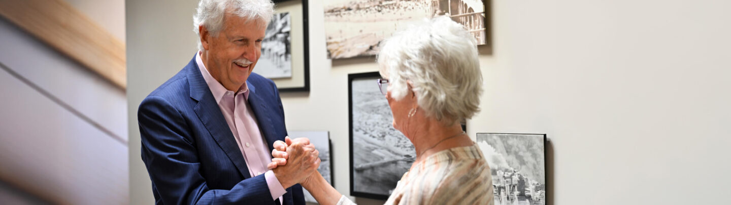 An older man in a blue suit smiling warmly and holding hands with an older woman in a hallway decorated with black-and-white photographs.