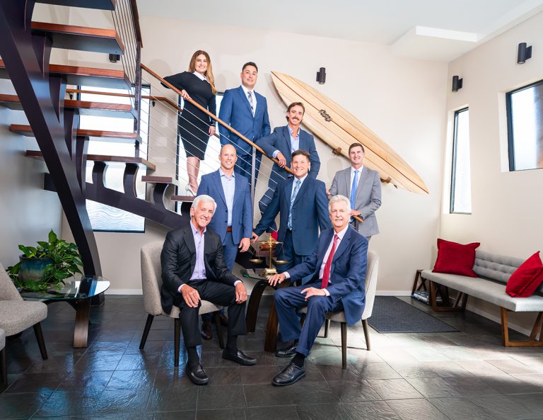 Seven professionally dressed people pose together in a modern office space with a staircase, indoor plants, a surfboard mounted on the wall, and seating with red pillows.