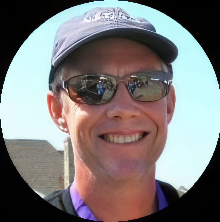 A smiling person wearing a dark baseball cap, sunglasses, and a purple shirt stands outdoors on a sunny day, with part of a building visible in the background. The photo is framed in a circular border.