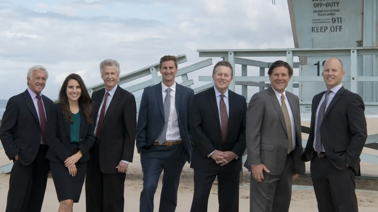 Seven professionally dressed people stand and smile on a sandy beach in front of a lifeguard tower, under a mostly cloudy sky.