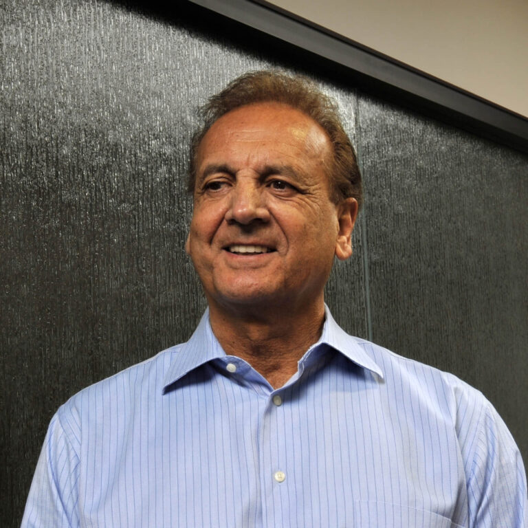 A middle-aged man with short brown hair is smiling while wearing a light blue, collared, button-down shirt. He is standing in front of a dark, textured wall.