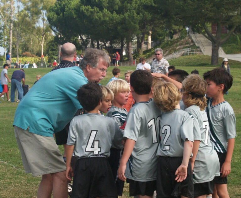 A coach in a teal shirt and khaki shorts huddles with a group of young boys in silver soccer uniforms on a grassy field, giving instructions before a game. Trees and spectators are visible in the background.