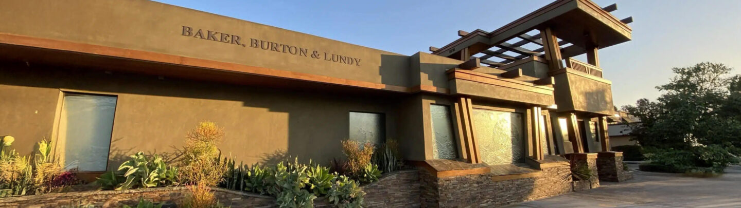 A modern building with Baker, Burton & Lundy written on the facade, large windows, brown and green exterior, and landscaped plants in front, photographed in warm sunlight.