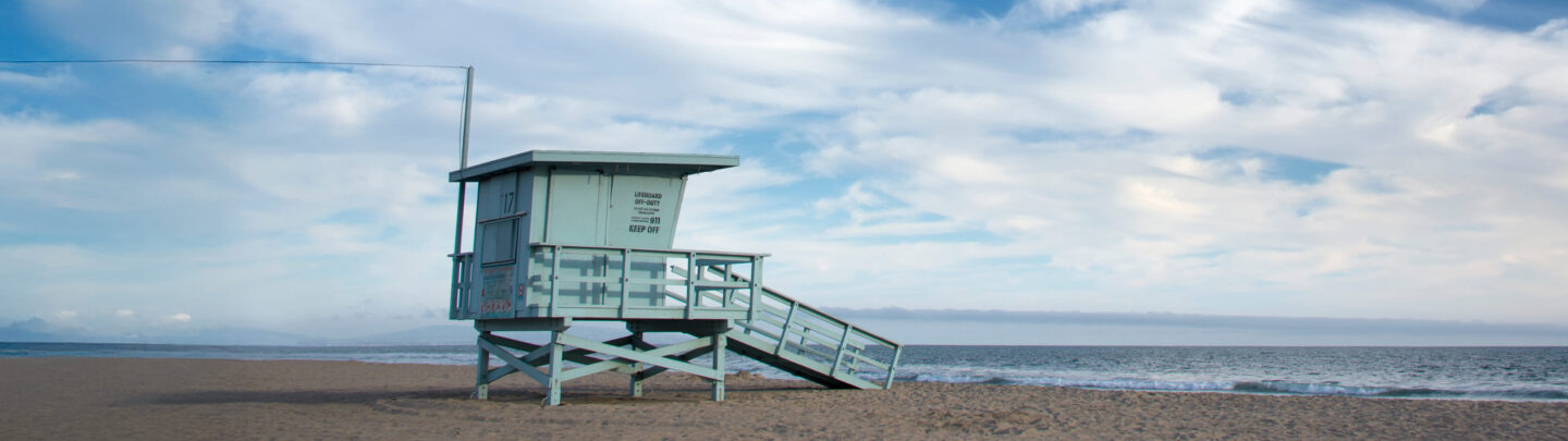 A light blue lifeguard tower stands on a sandy beach under a cloudy sky, with the ocean visible to the right and no people in sight.