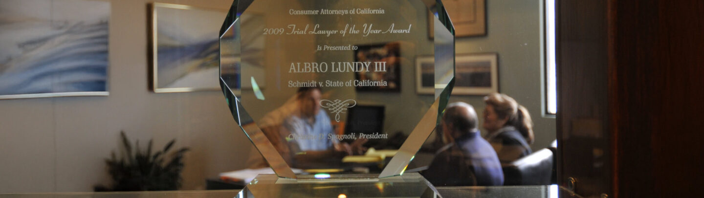 A clear glass award with engraved text sits on a desk. In the background, three people are having a meeting in an office decorated with framed pictures and plants.