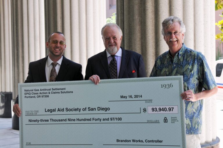 Three men stand outside holding a large ceremonial check for $93,940.97 made out to the Legal Aid Society of San Diego, dated May 16, 2014, from the Natural Gas Antitrust Settlement.