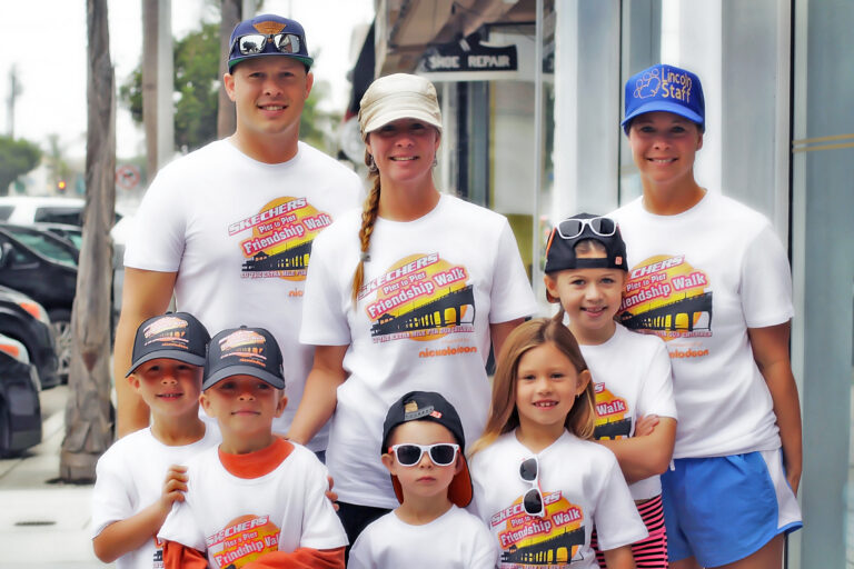 A group of adults and children wearing matching “Friendship Walk” T-shirts and hats pose together and smile for the camera on a city sidewalk.