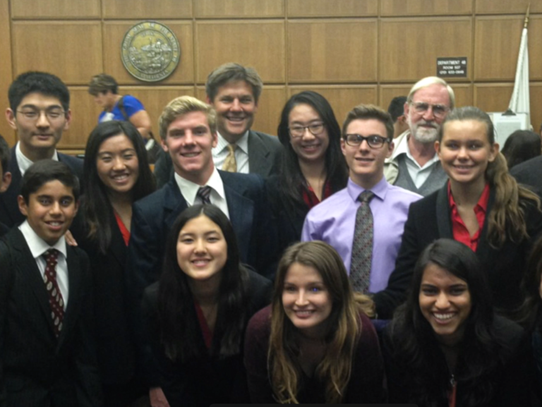 A group of twelve people, mostly young adults in business attire, smiling and posing in a wood-paneled room with a seal and a flag visible in the background, suggesting a formal or official setting.