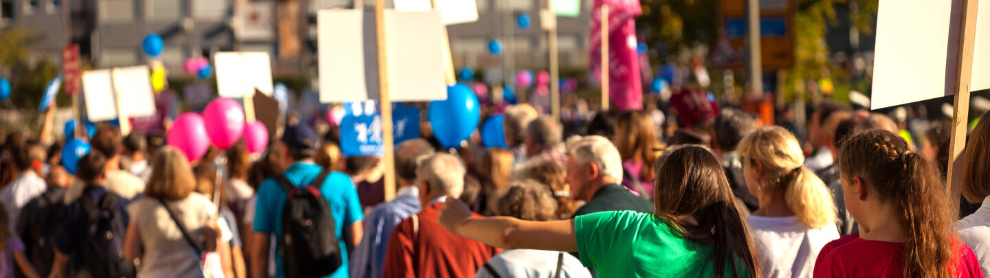 A large group of people march outdoors on a sunny day, holding blank signs and balloons, participating in a protest or demonstration. The crowd moves along a city street, with buildings and trees in the background.