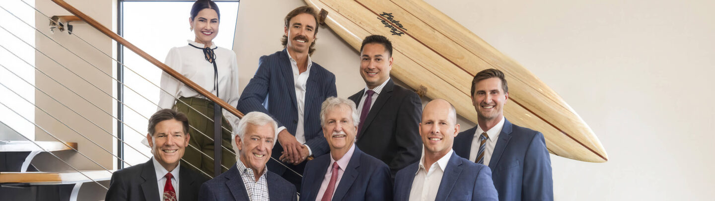 Eight professionally dressed people, seven men and one woman, pose and smile together in an office setting near a staircase with a large surfboard mounted on the wall behind them.