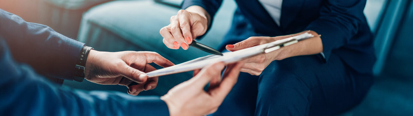 Two people in business attire sit together, one holding a clipboard with papers while the other points at it with a pen, suggesting a discussion or review of documents. Only their hands and part of their bodies are visible.