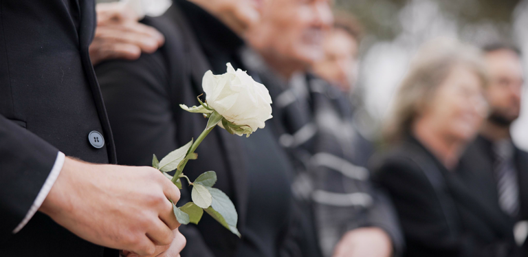 A person in a suit holds a white rose, standing among a group of people dressed in black, likely attending a funeral or memorial service. The mood appears somber and respectful.