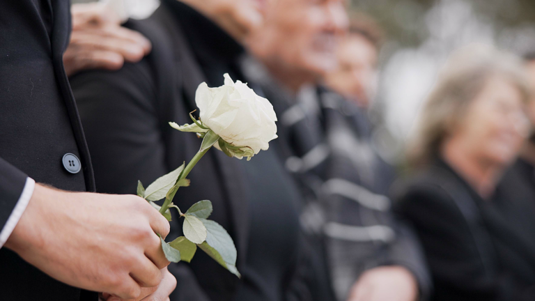 A person in a suit holds a white rose, standing among a group of people dressed in black, likely attending a funeral or memorial service. The mood appears somber and respectful.