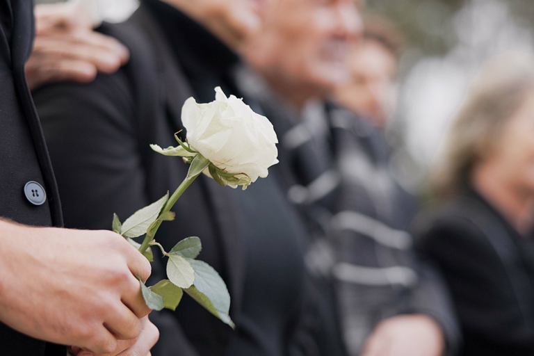 A person in a suit holds a white rose, standing among a group of people dressed in black, likely attending a funeral or memorial service. The mood appears somber and respectful.