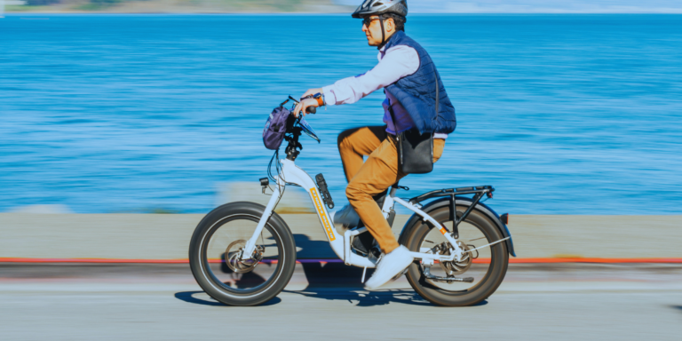 A person wearing a helmet, sunglasses, blue vest, and brown pants rides an electric bicycle along a waterfront road with blue water in the background.
